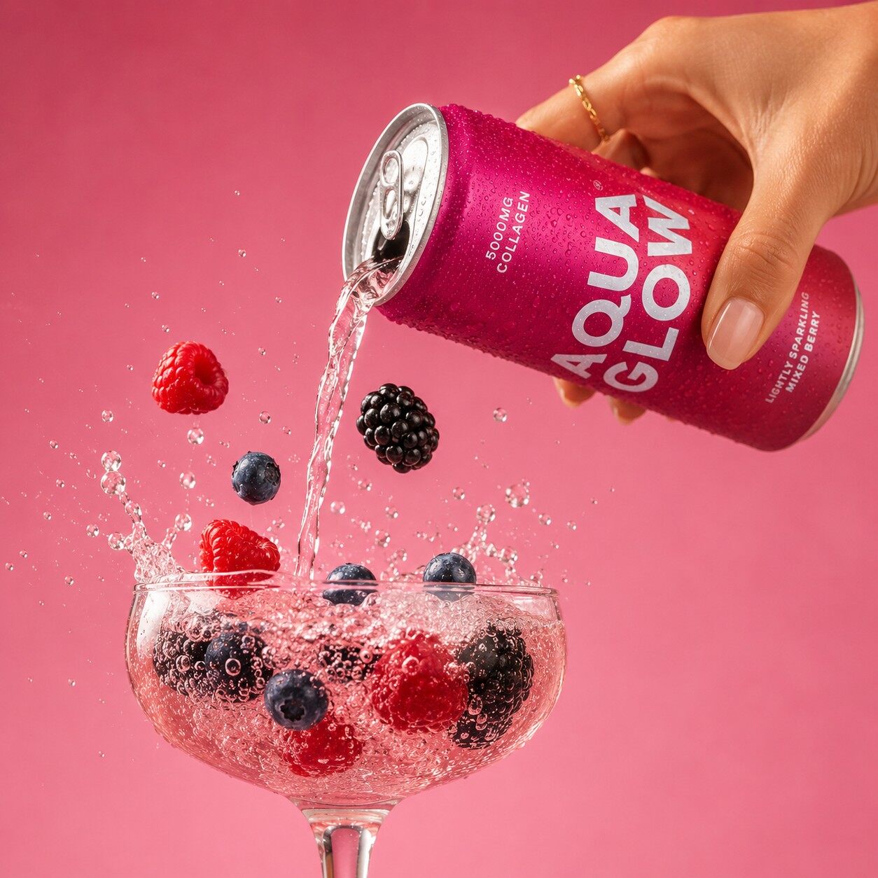 A hand pouring a pink can of Aqua Glow Berry Crush into a coupe glass with raspberries and blackberries