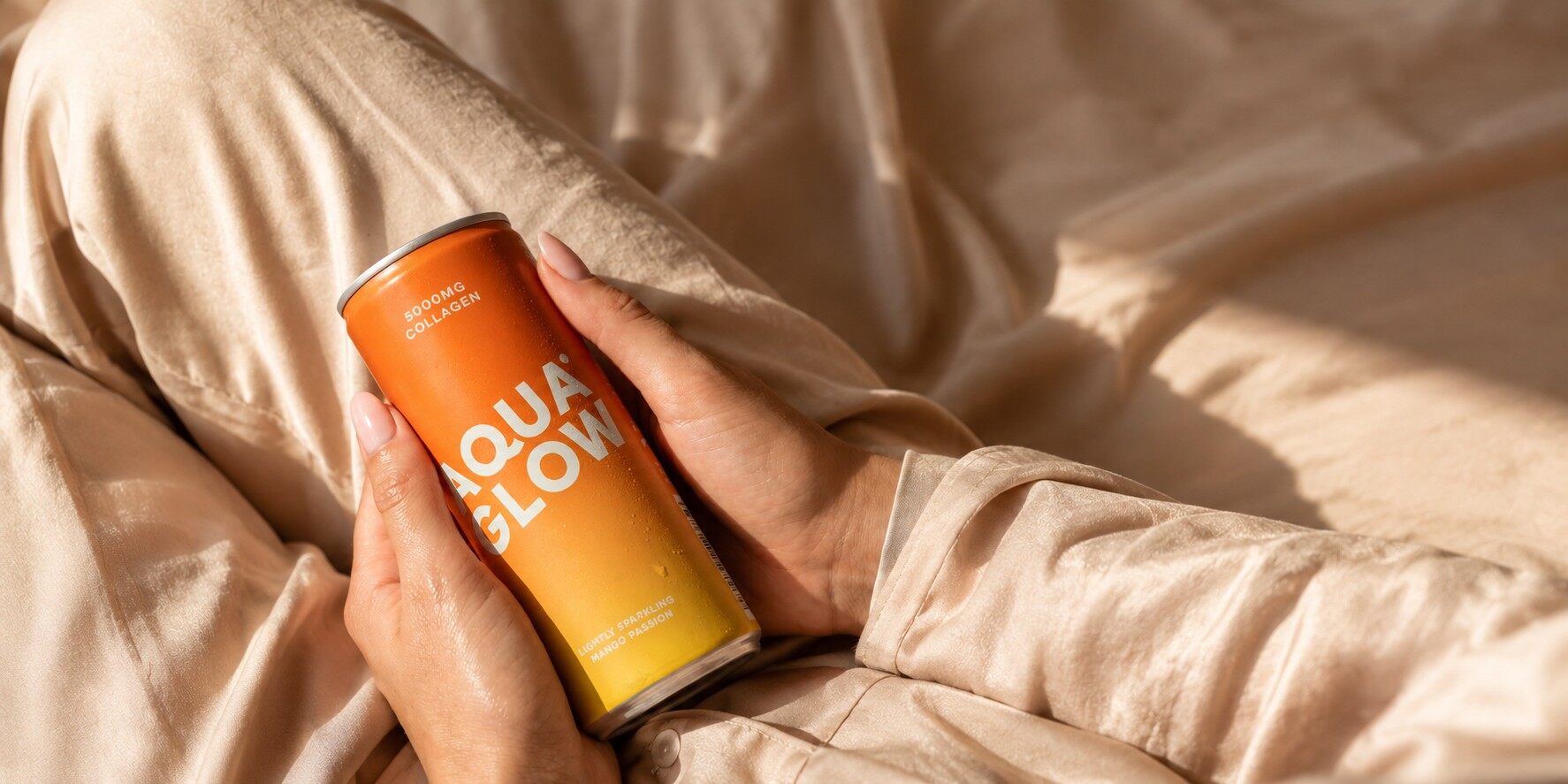Two hands holding an orange can of Aqua Glow Mango Passion against beige satin bedding