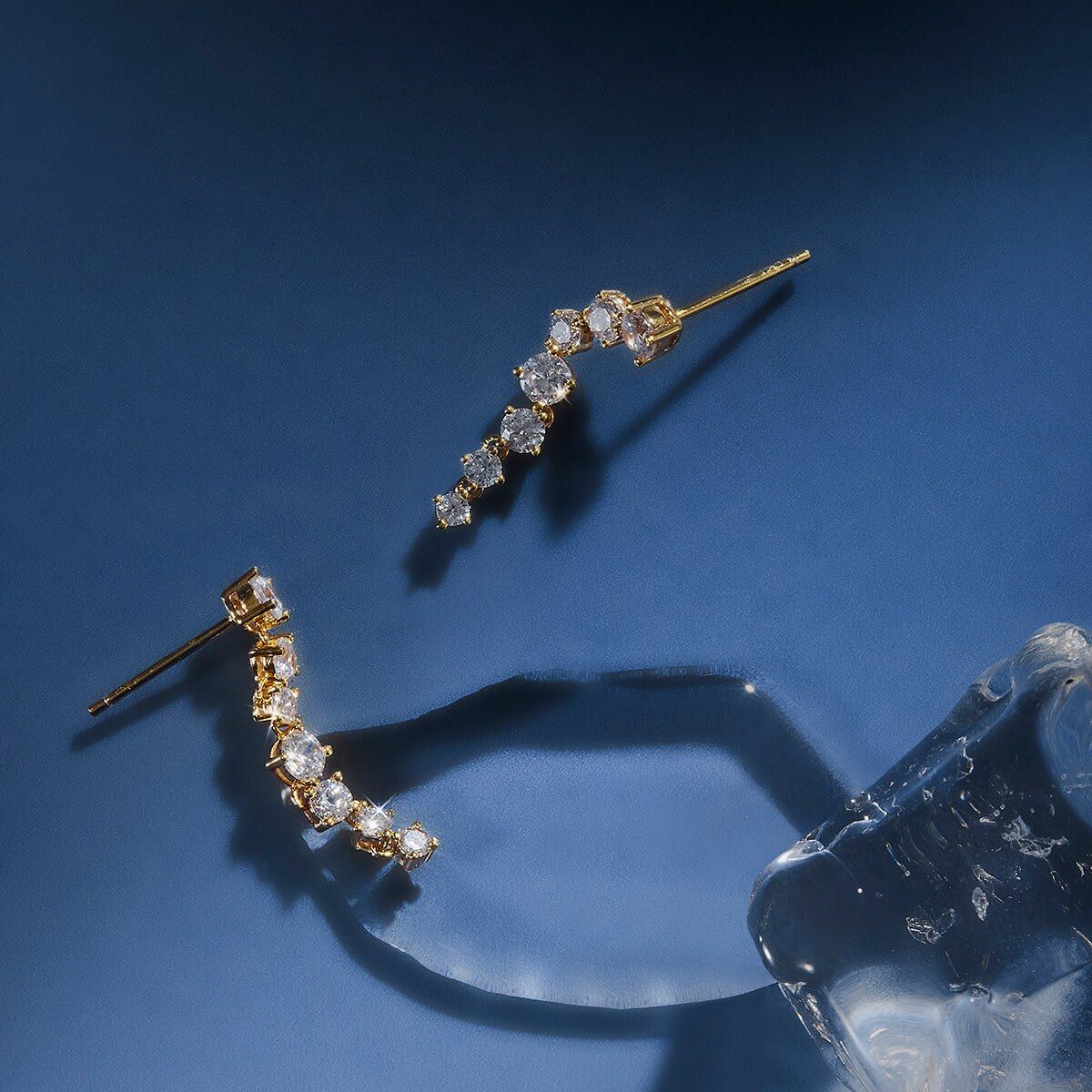 Two Astrid & Miyu gold ear cuffs set with crystals, photographed on a deep blue background