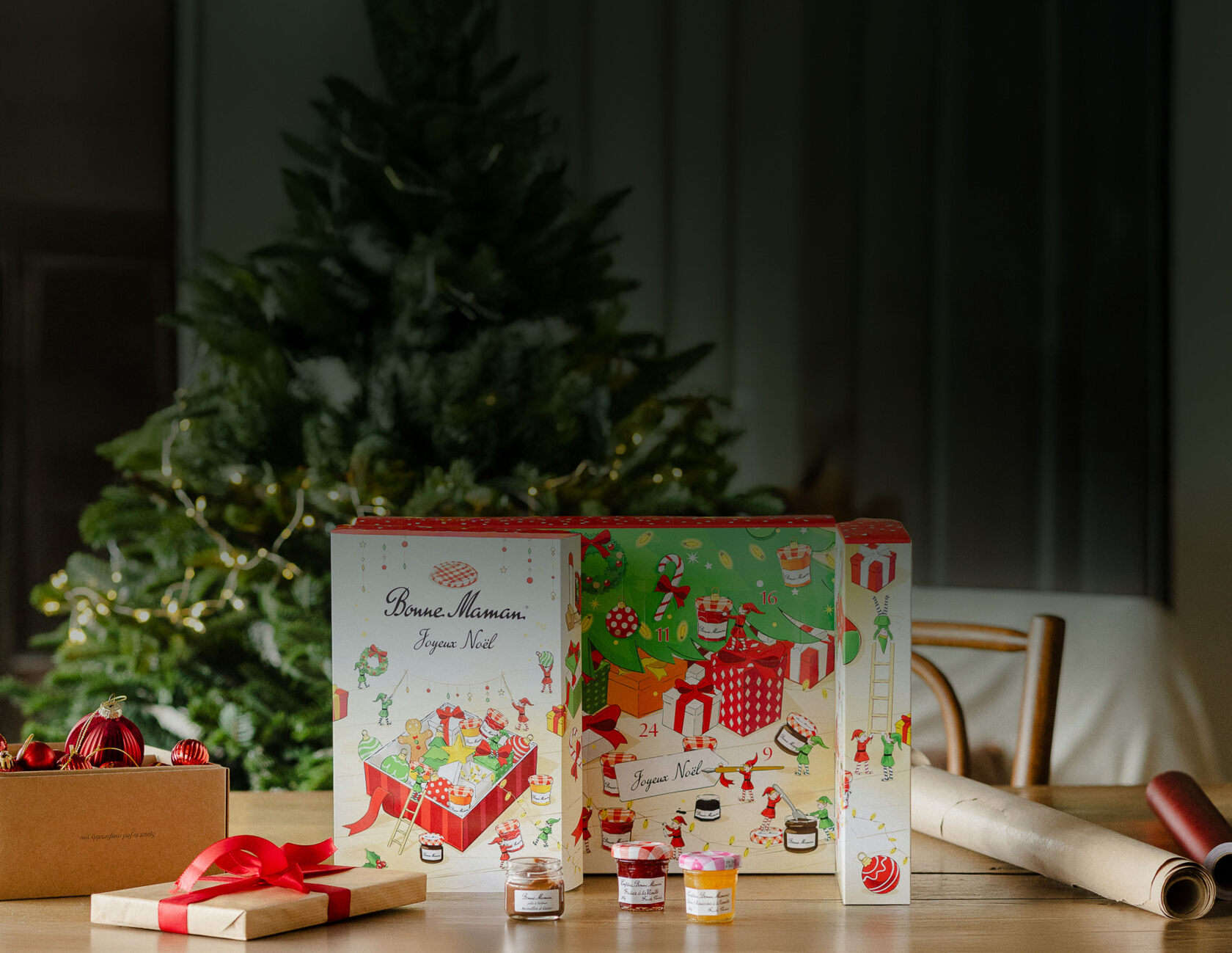The Bonne Maman advent calendar 2026 standing open on a wooden table in front of a Christmas tree, with mini jars and wrapped gifts