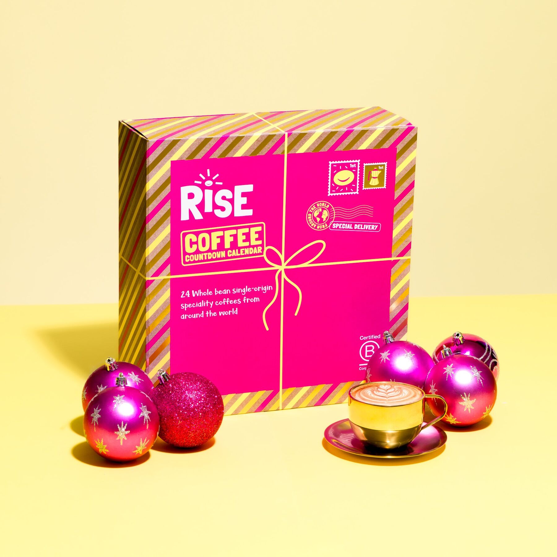 The pink and yellow RISE coffee advent calendar box tied with a ribbon, with pink baubles and a cup of coffee on a yellow background