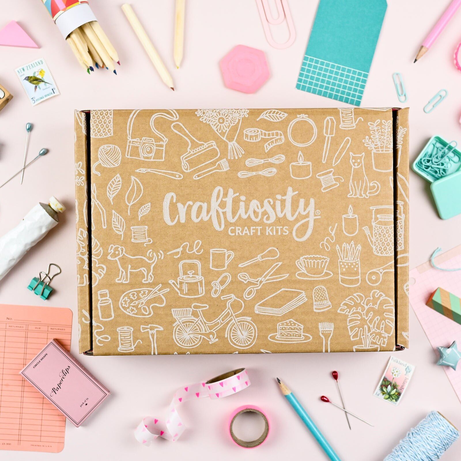 A closed Craftiosity craft kit box printed with white illustrations, surrounded by pencils, washi tape, pins and paper