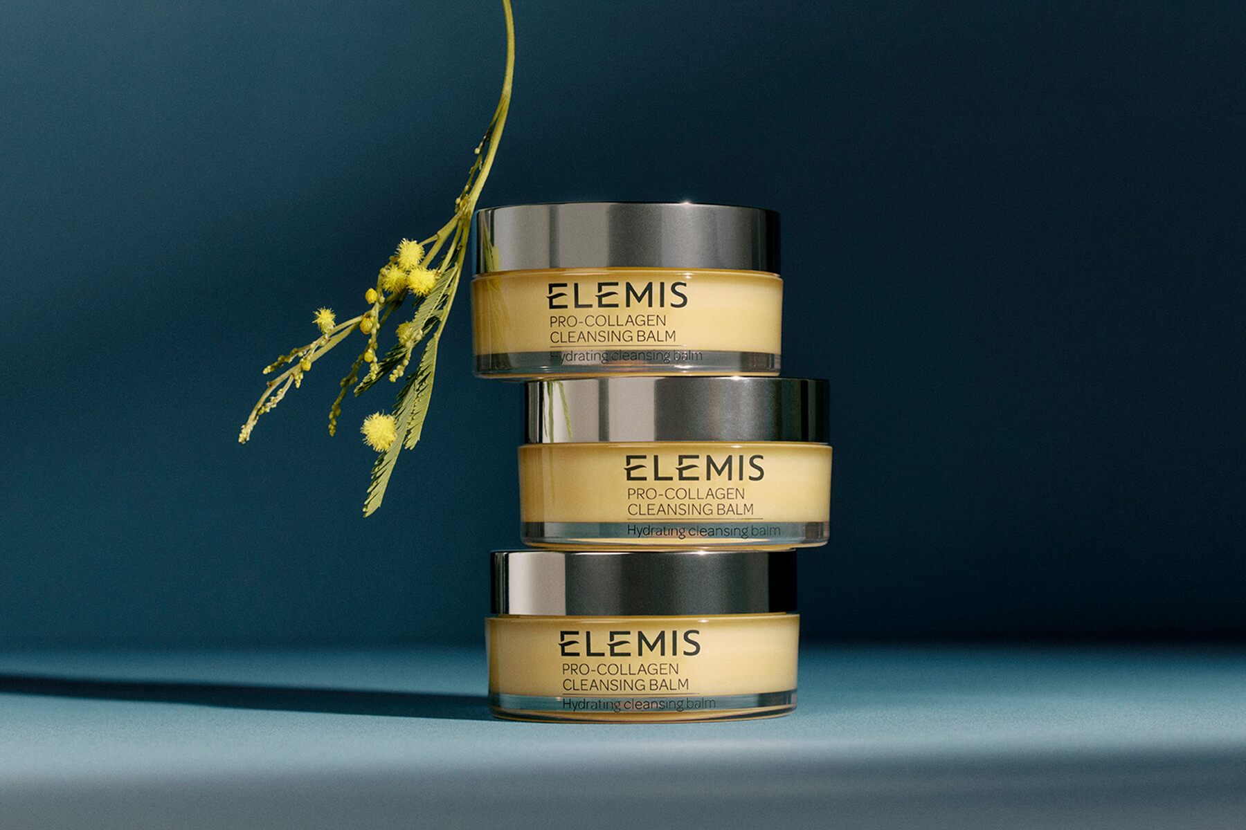 Three jars of ELEMIS Pro-Collagen Cleansing Balm stacked on a dark blue background with a sprig of yellow mimosa