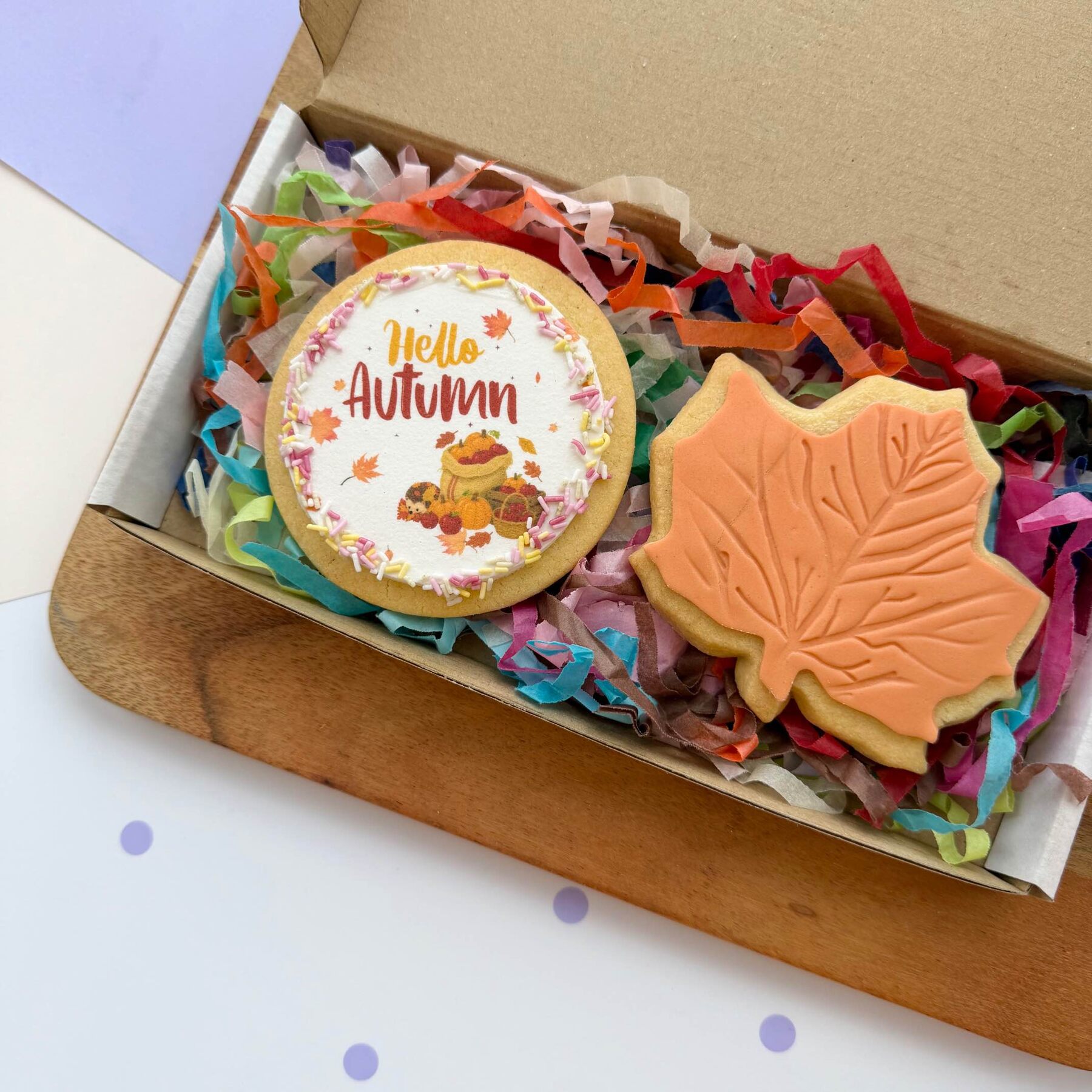 A Hello Autumn cookie and an orange maple leaf cookie in a gift box with rainbow tissue
