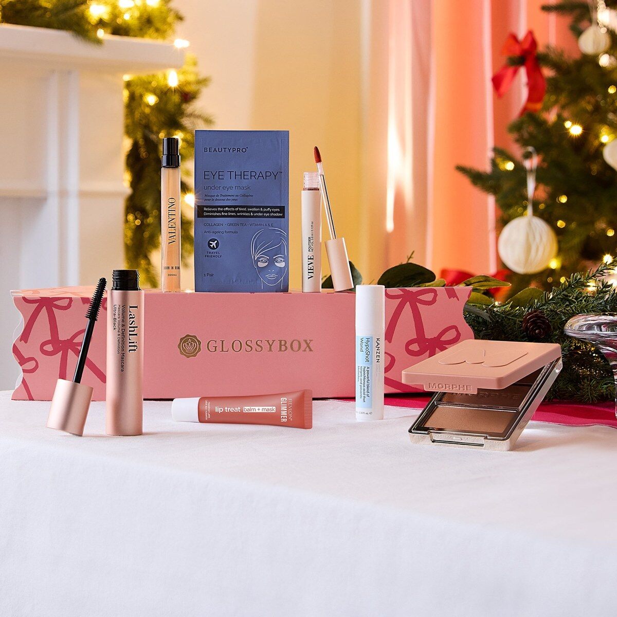 The pink GLOSSYBOX Christmas Cracker 2026 on a festive table with a mascara, a Valentino perfume, an eye mask and a face palette