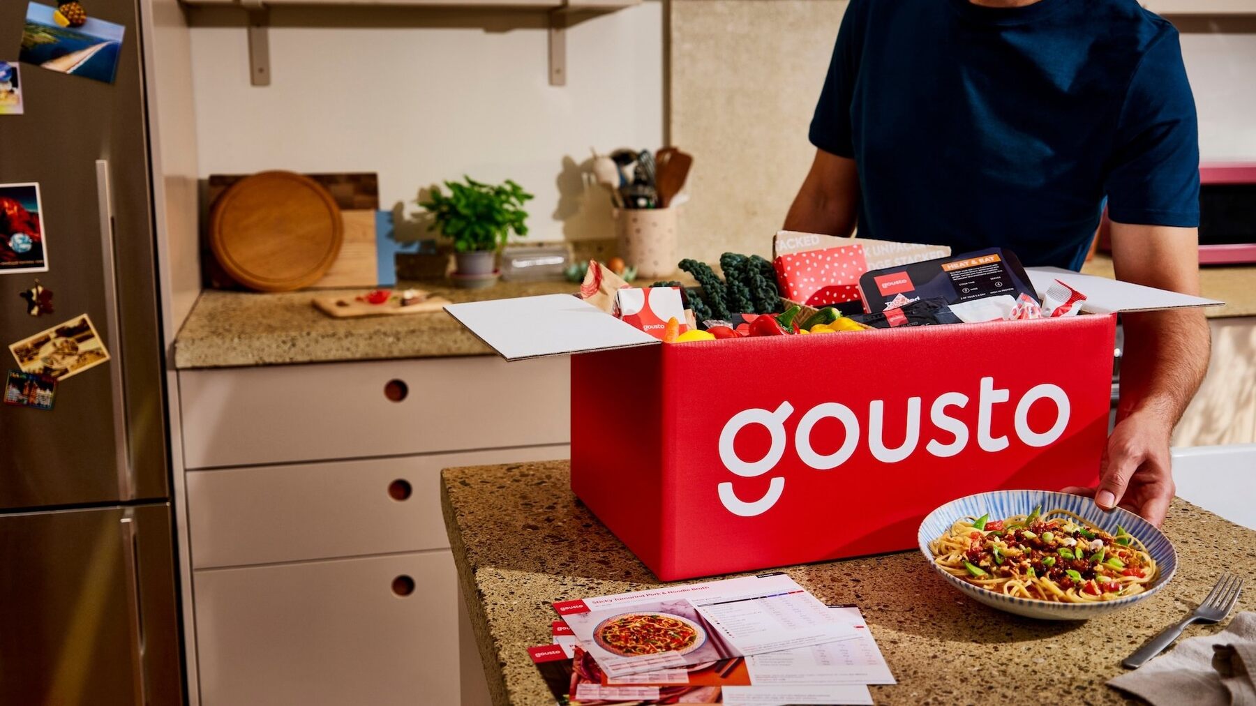 A man unpacking a red Gousto recipe box in a home kitchen next to a bowl of pasta