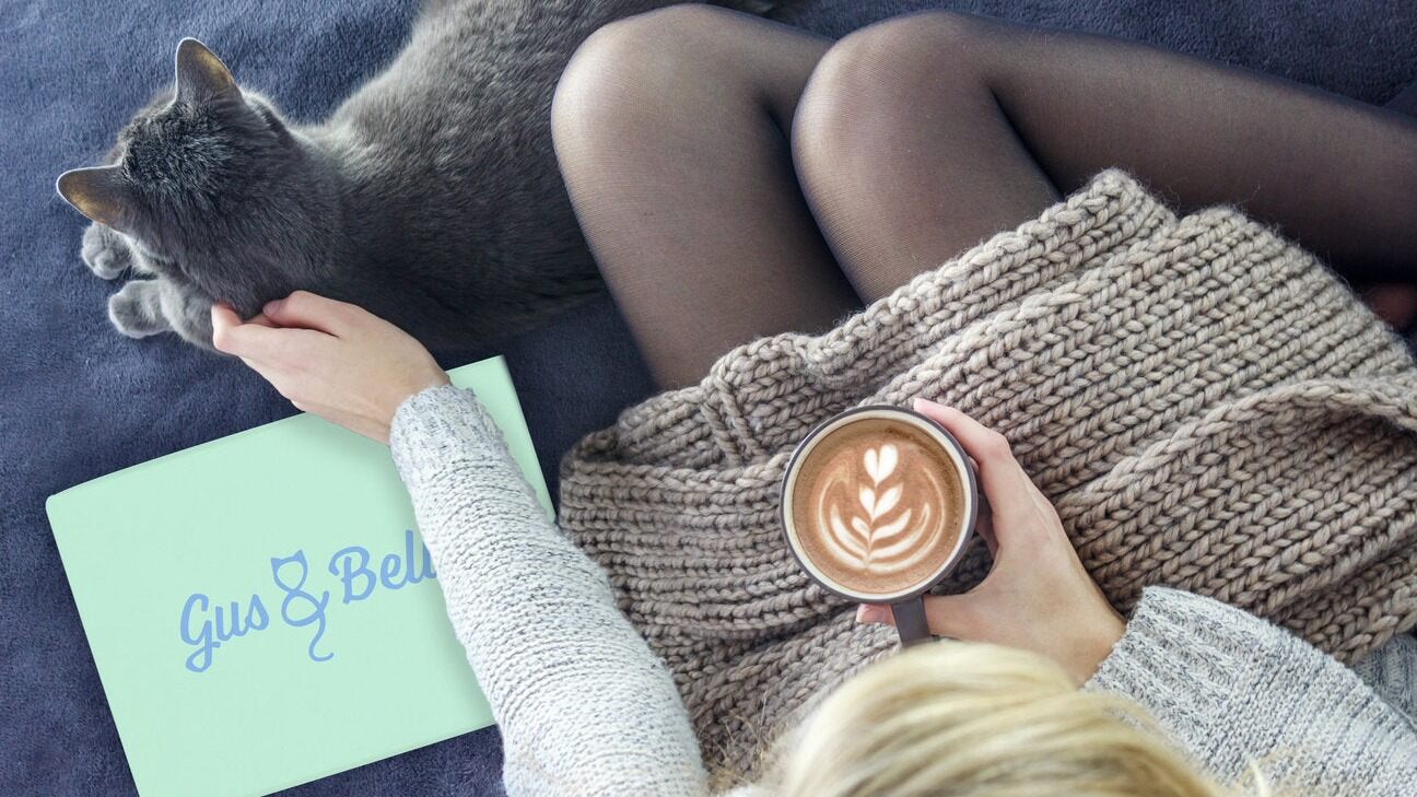 A grey cat on a woman's lap in a cosy jumper, with a latte and a Gus & Bella box beside her