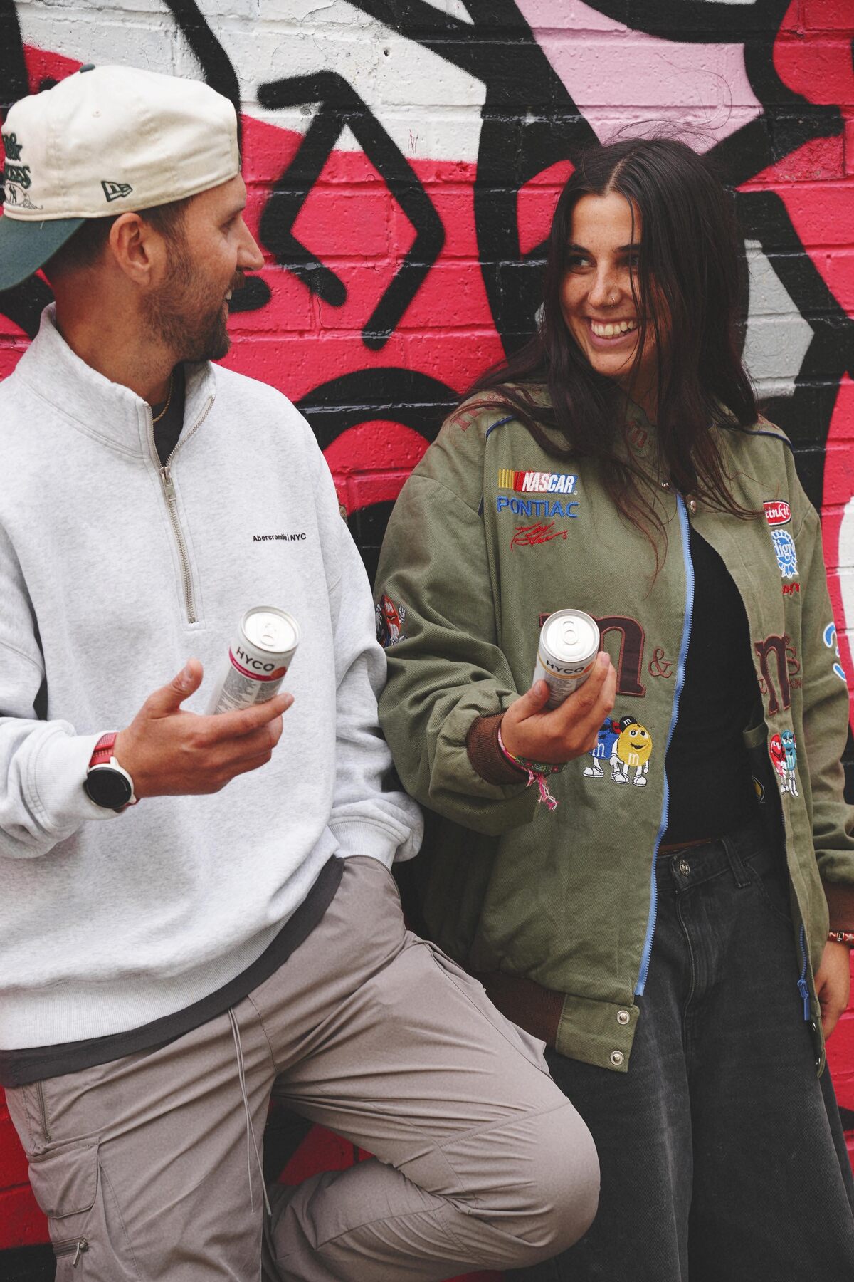 A man and a woman laughing in a city street, each holding a can of HYCO
