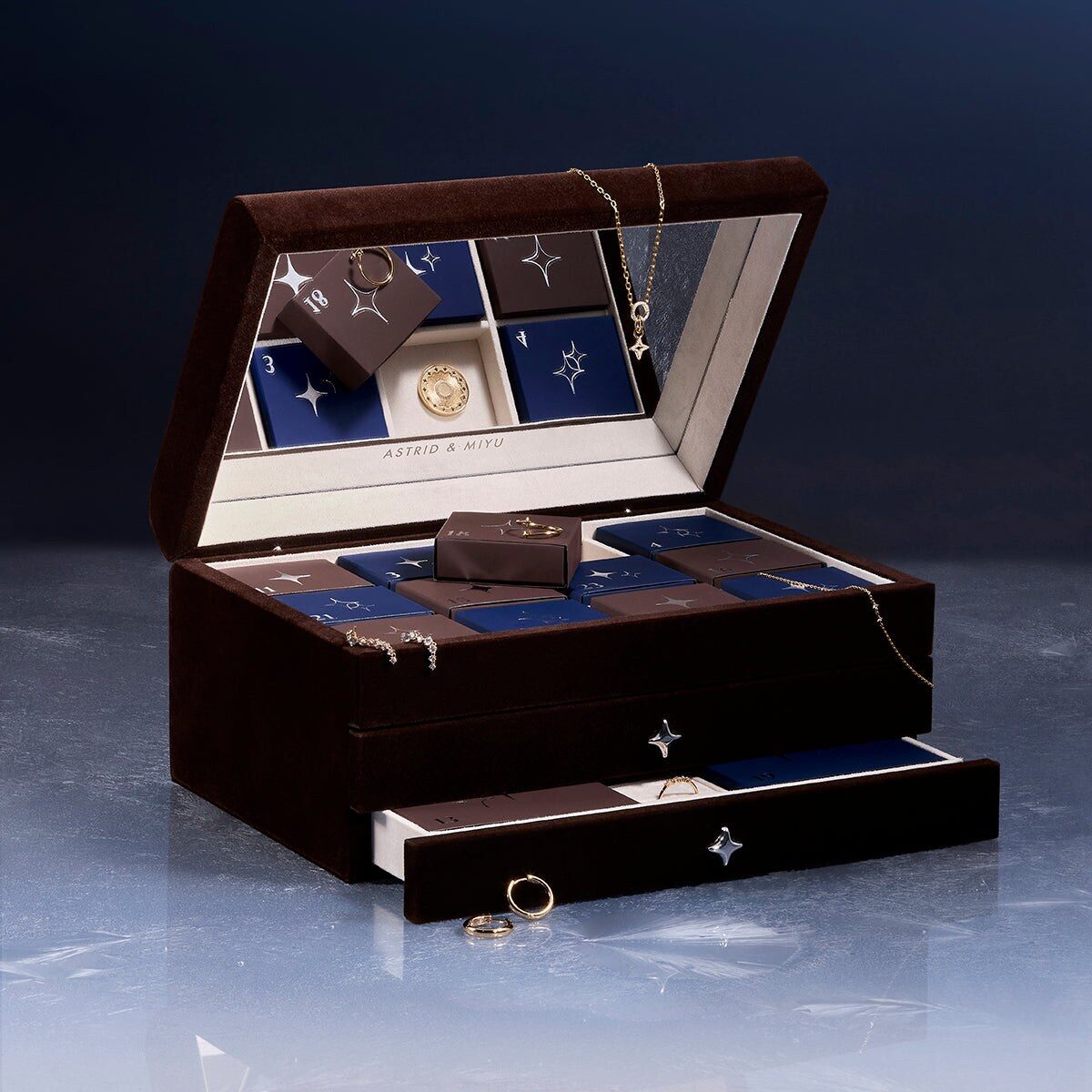 The Astrid & Miyu 24 Day advent calendar in gold, an open dark wood jewellery box with navy star drawers, on a frosted surface