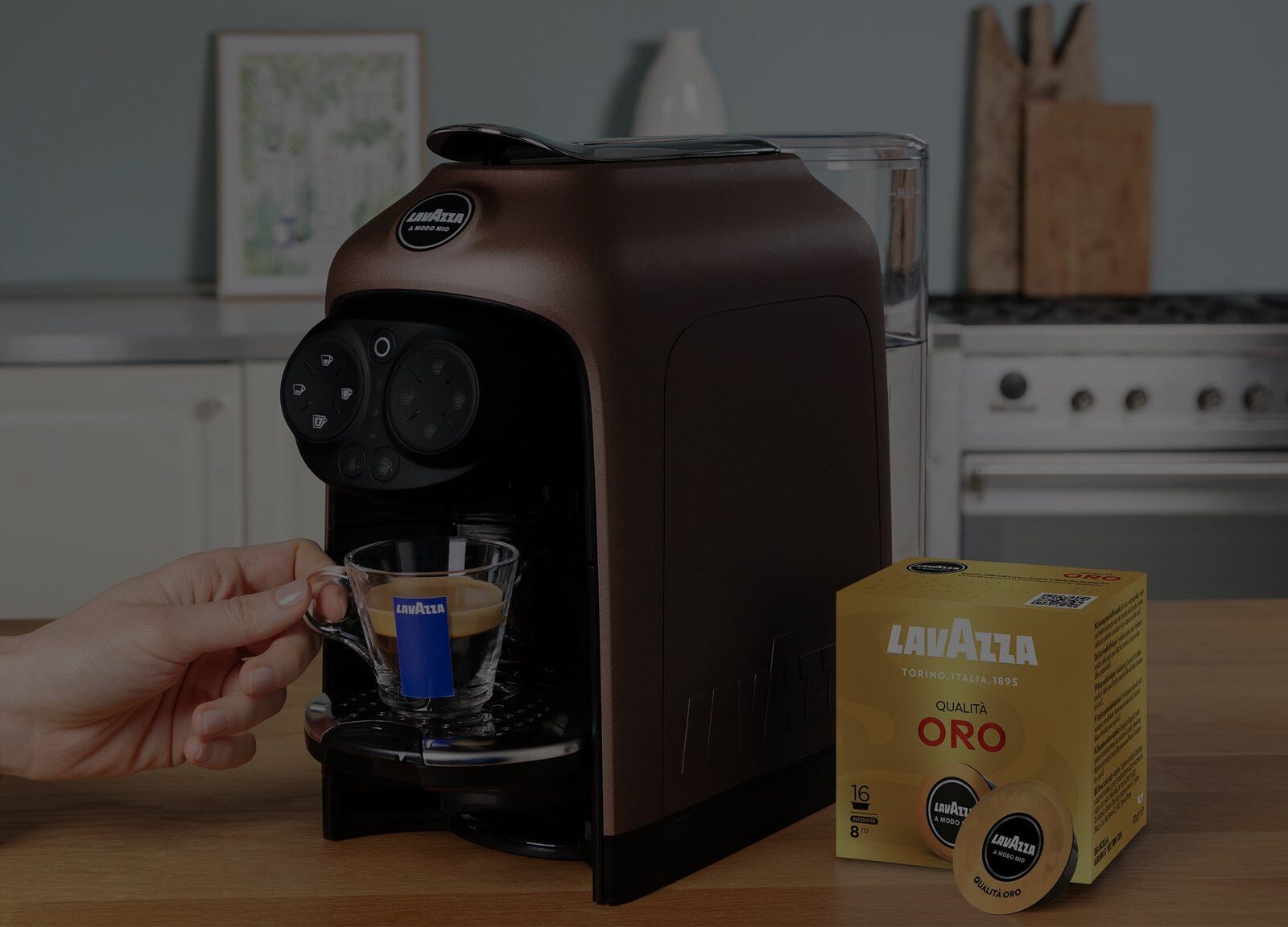 Hand taking an espresso cup from a Lavazza A Modo Mio machine beside a box of capsules