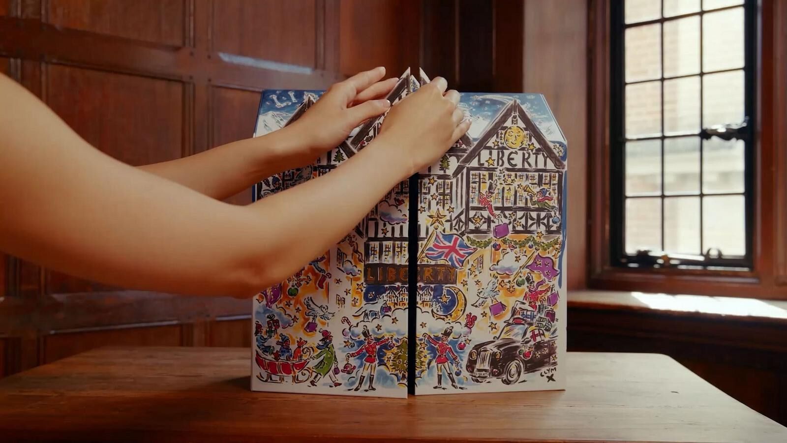 A hand opening the Liberty beauty advent calendar, shaped like the Liberty Tudor building, on a wooden table