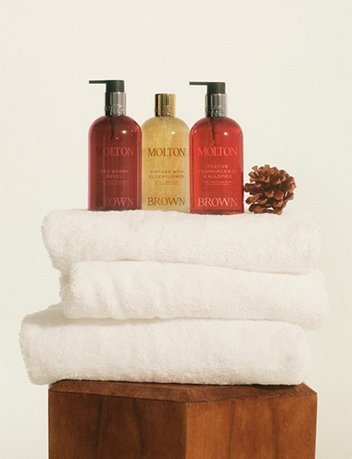 Three Molton Brown pump bottles in red and gold standing on a stack of white towels next to a pine cone