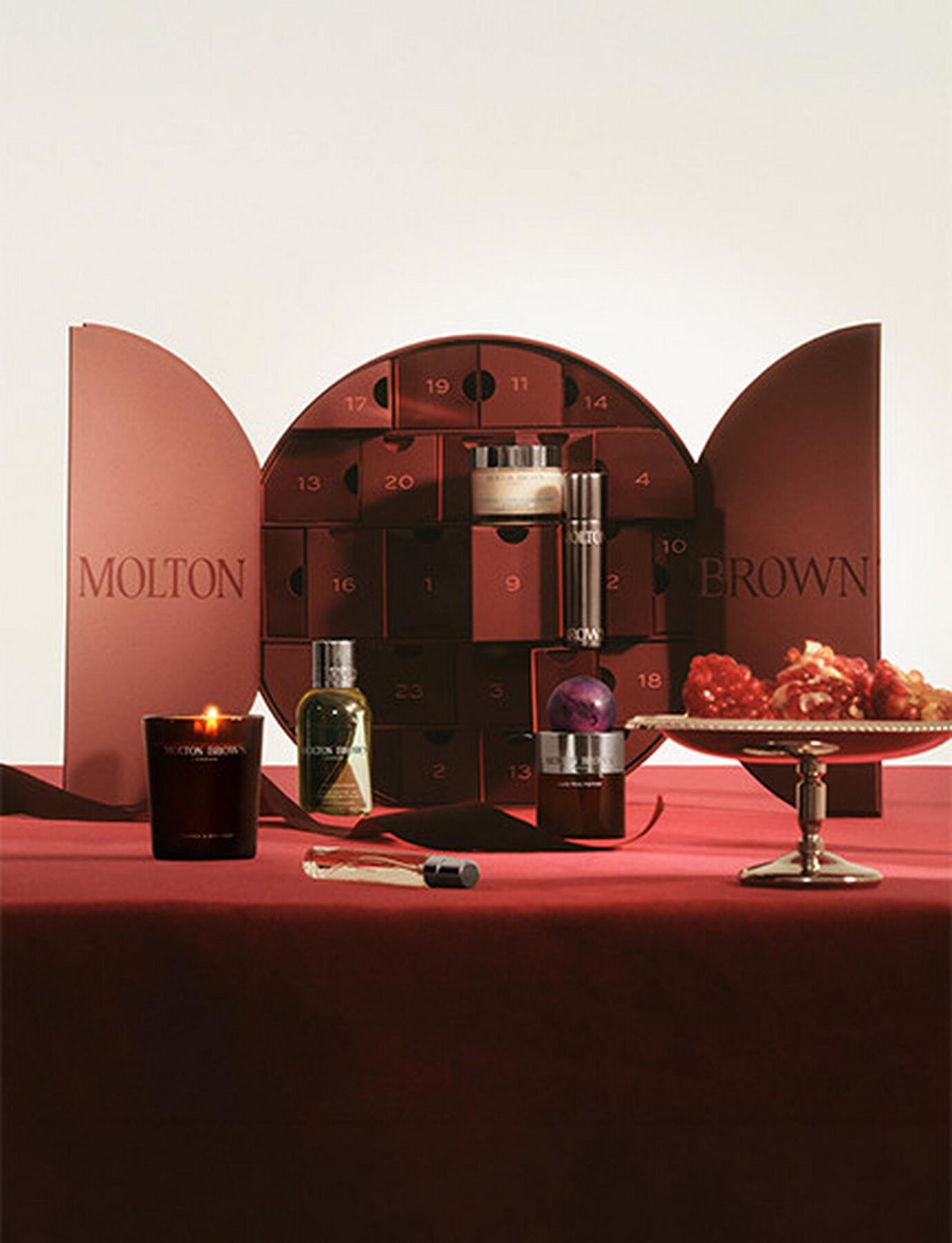 The open Molton Brown advent calendar 2026 on a red tablecloth with a candle, a fragrance bottle and a bowl of pomegranates