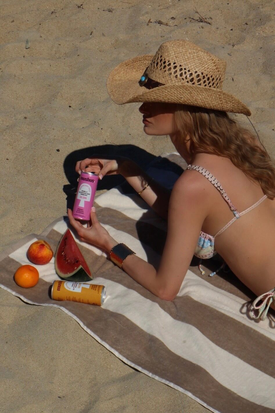 A woman lying on a striped beach towel on the sand, holding a pink can of Mushus Raspberry Fizz
