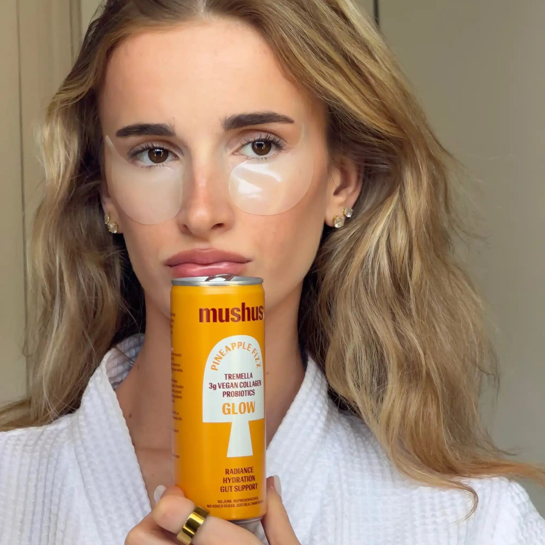 A Mushus Pineapple Fizz can held up by a woman in a white robe wearing under-eye patches
