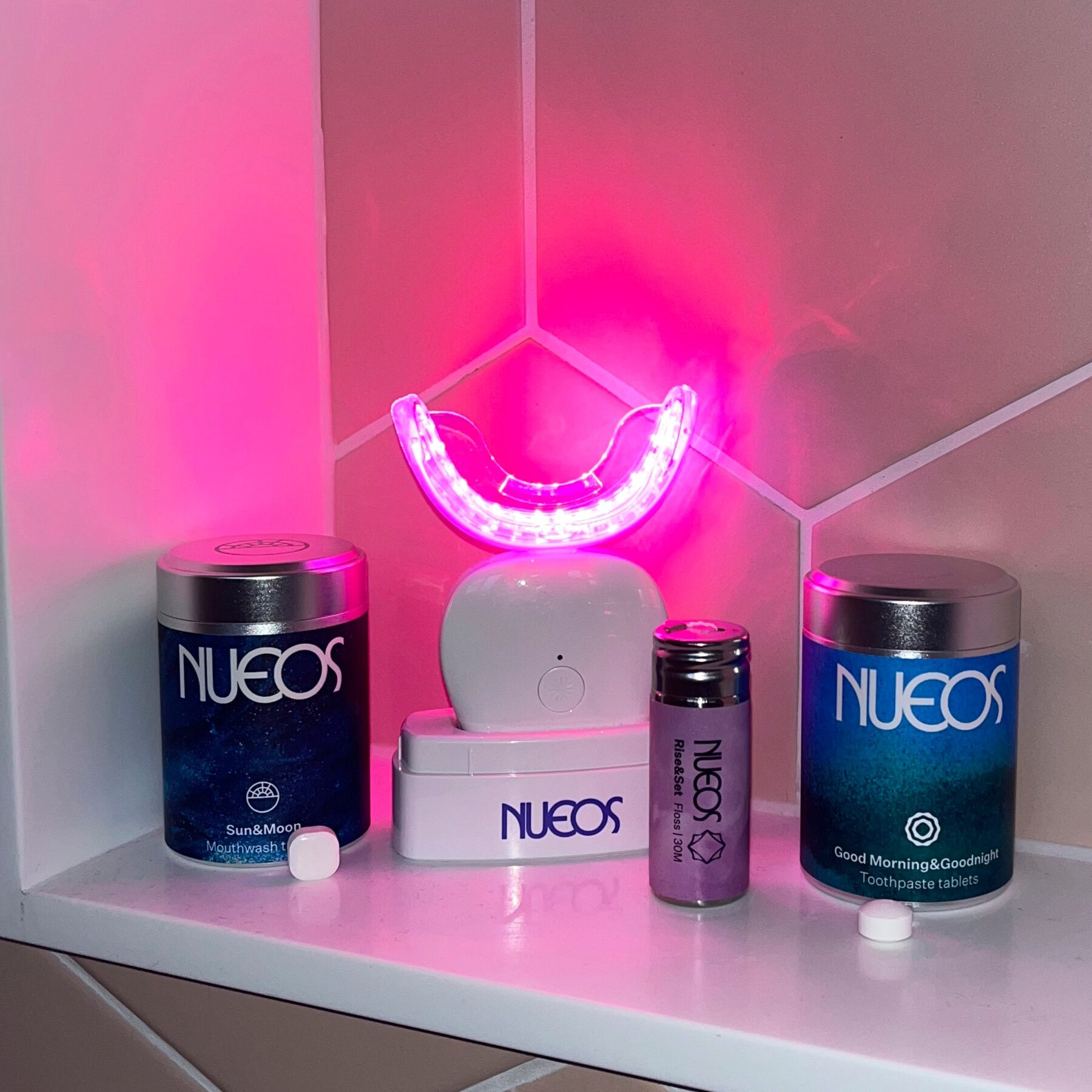 The NUEOS Light Bath glowing pink on a bathroom shelf beside the blue and green NUEOS storage tins