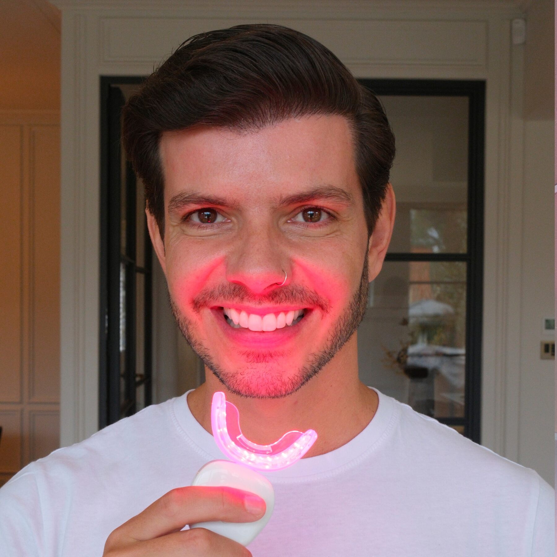 A smiling man in a white T-shirt holding the glowing NUEOS Light Bath mouthpiece in his mouth at home