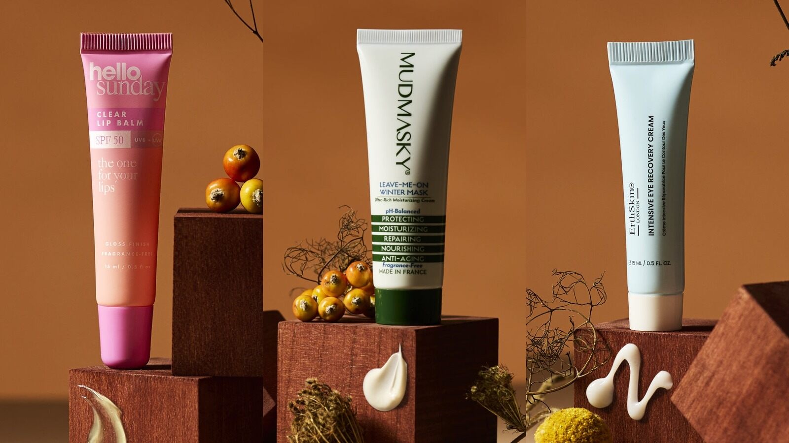The Hello Sunday SPF50 lip balm, MUDMASKY Leave-Me-On Winter Mask and Erth Skin London eye cream on wooden stands with autumn berries