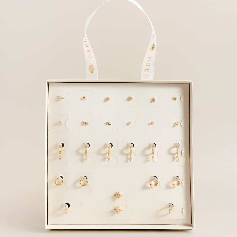 The Orelia 25 Days of Ear Stacking advent calendar open on a cream background, showing gold hoops and studs in rows