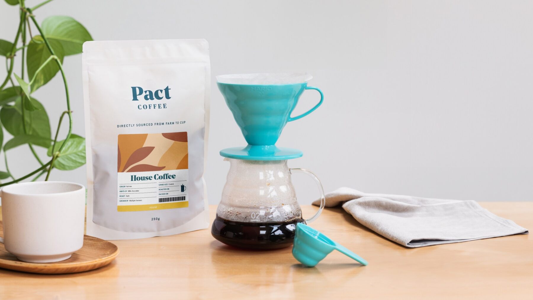 A bag of Pact House Coffee next to a pour-over brewer and a mug on a wooden table