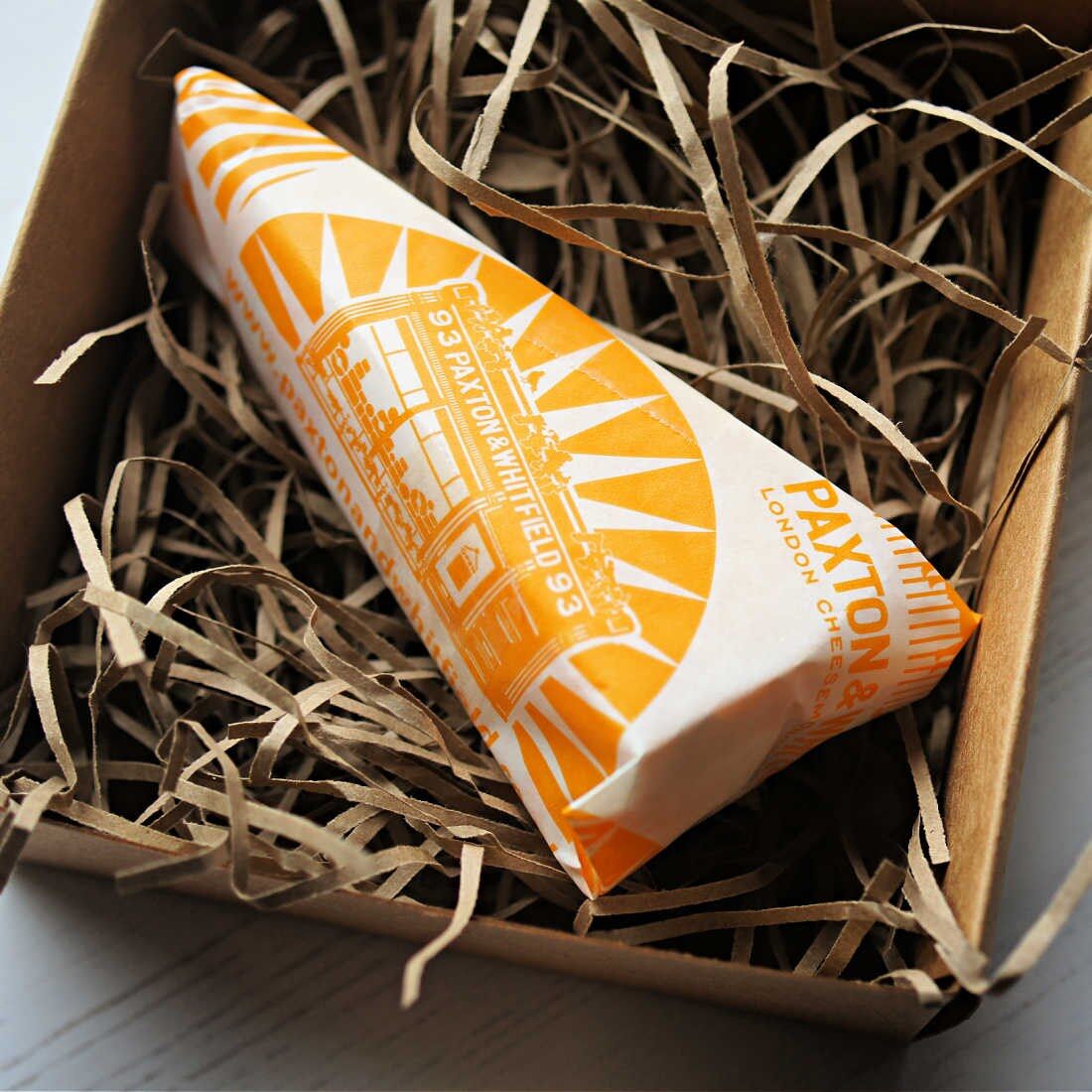 A wedge of cheese wrapped in Paxton and Whitfield paper in a box lined with straw