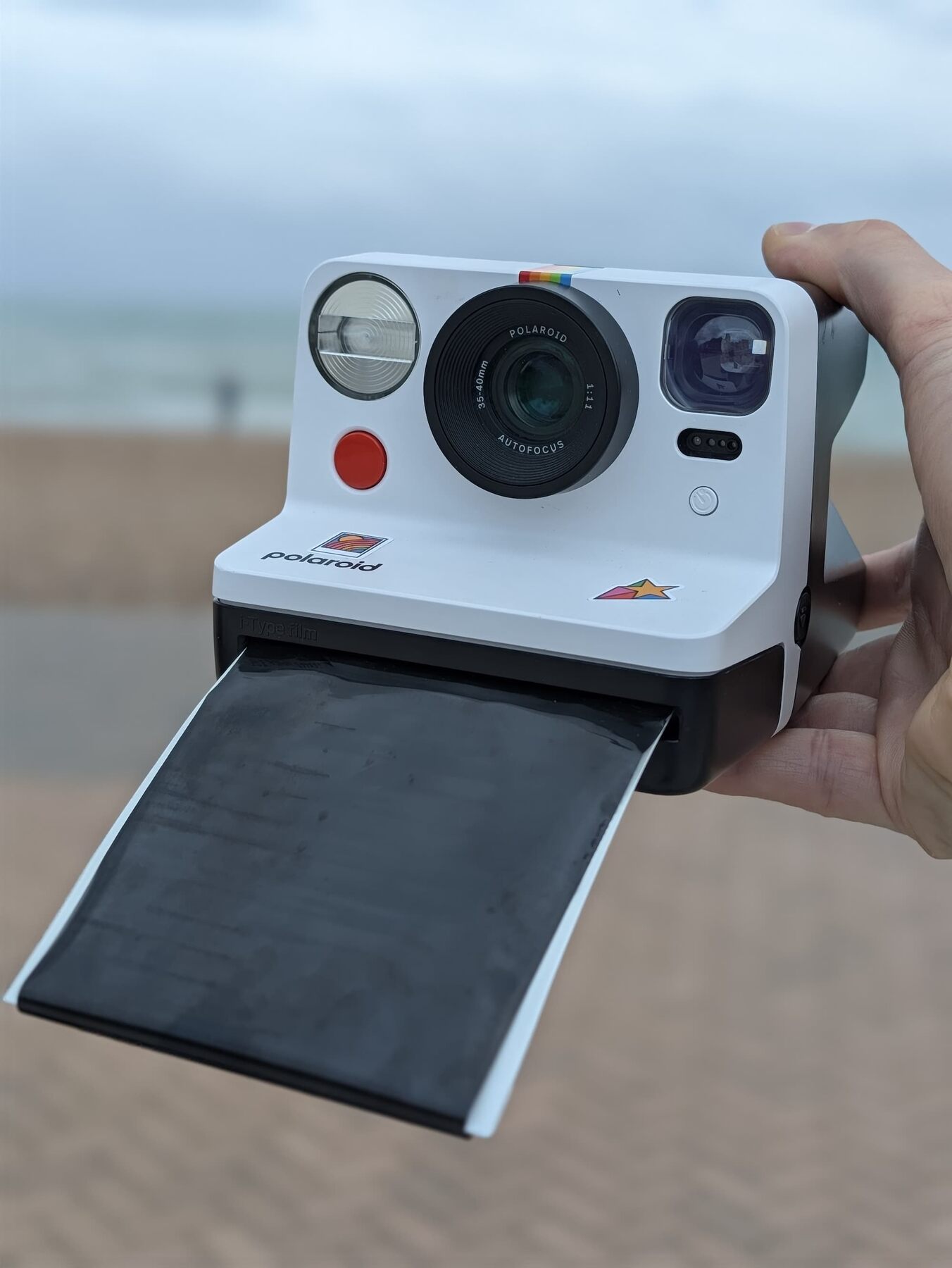 Hand holding a white Polaroid Now instant camera on a seafront with a grey sea behind, a photo ejecting from the film slot