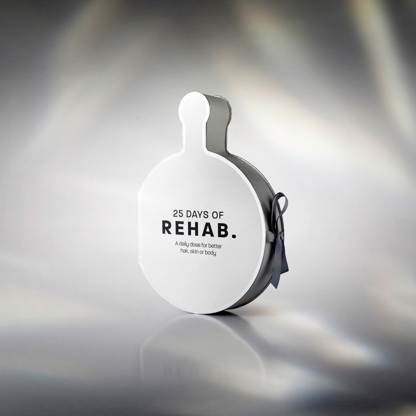 The REHAB. Dose advent calendar, a white box shaped like a hand mirror with a silver ribbon, on a shimmering grey background
