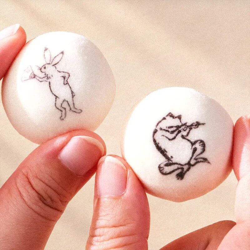 Two round white marshmallows printed with black ink drawings of a rabbit and a frog, held in a hand