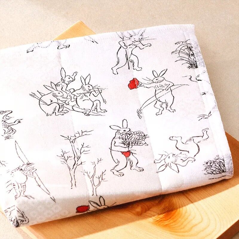 A white kaya cloth printed with black ink drawings of frogs, rabbits and monkeys, folded on a wooden table