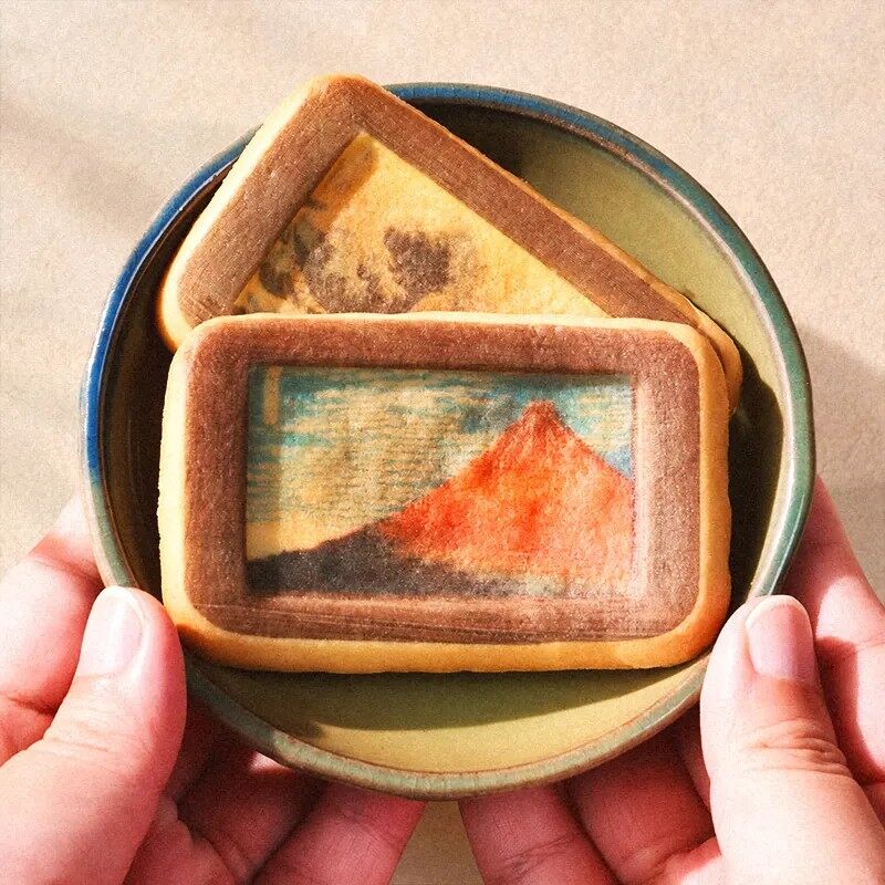 A hand holding a rectangular biscuit printed with a ukiyo-e style picture of a red Mount Fuji