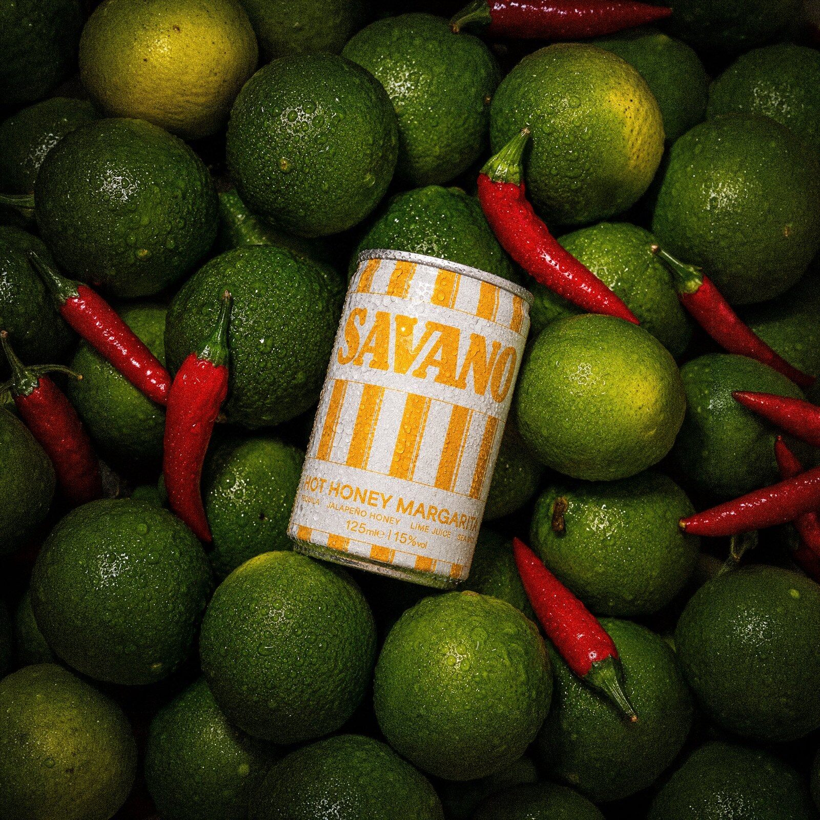 A striped orange can of Savano Hot Honey Margarita lying on a bed of limes and red chillies
