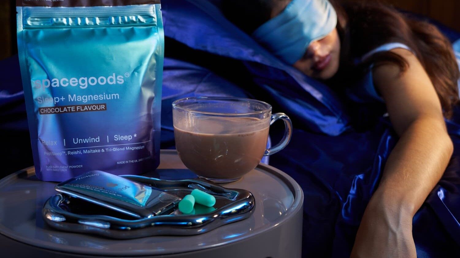 A pouch of Spacegoods Sleep+ and a mug of hot chocolate on a bedside table while a woman sleeps in an eye mask