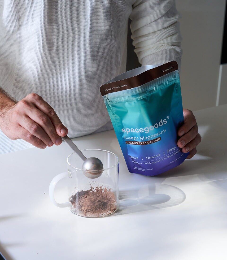 A hand adding a scoop of Spacegoods Sleep+ powder to a glass mug next to the pouch