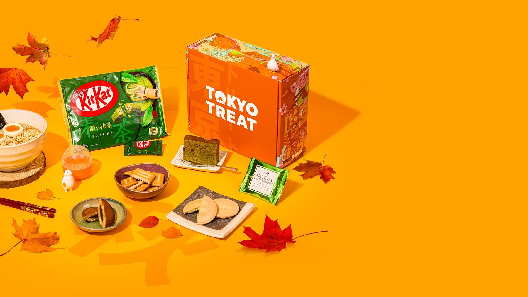 An orange TokyoTreat box with matcha KitKats, dorayaki and Japanese snacks among autumn leaves