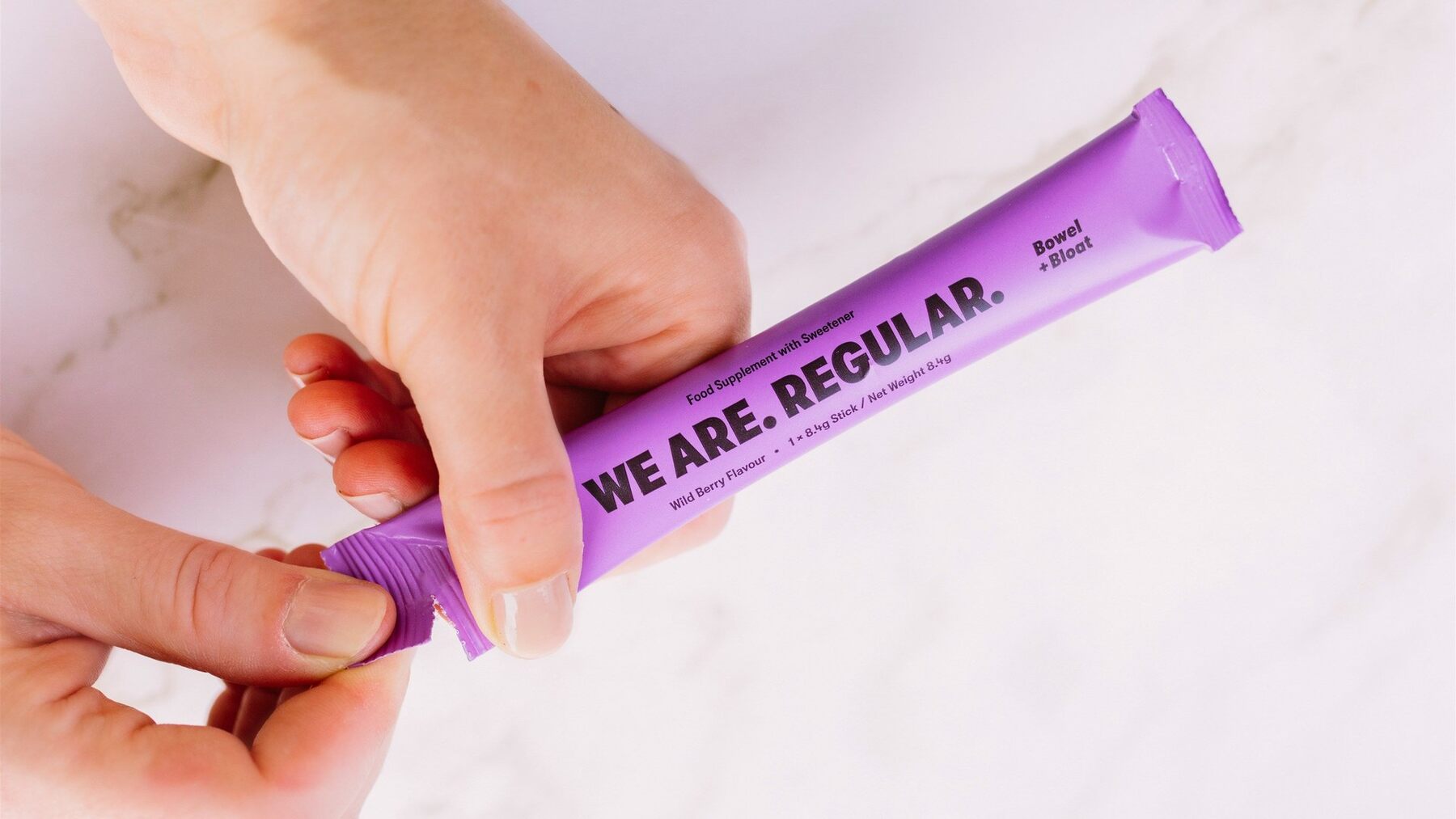 A hand tearing open a purple We Are Regular Bowel + Bloat stick on a white marble surface