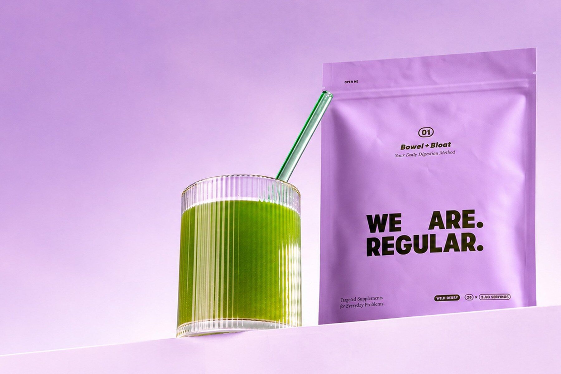 We Are Regular pouch next to a glass of the green drink on a lilac background