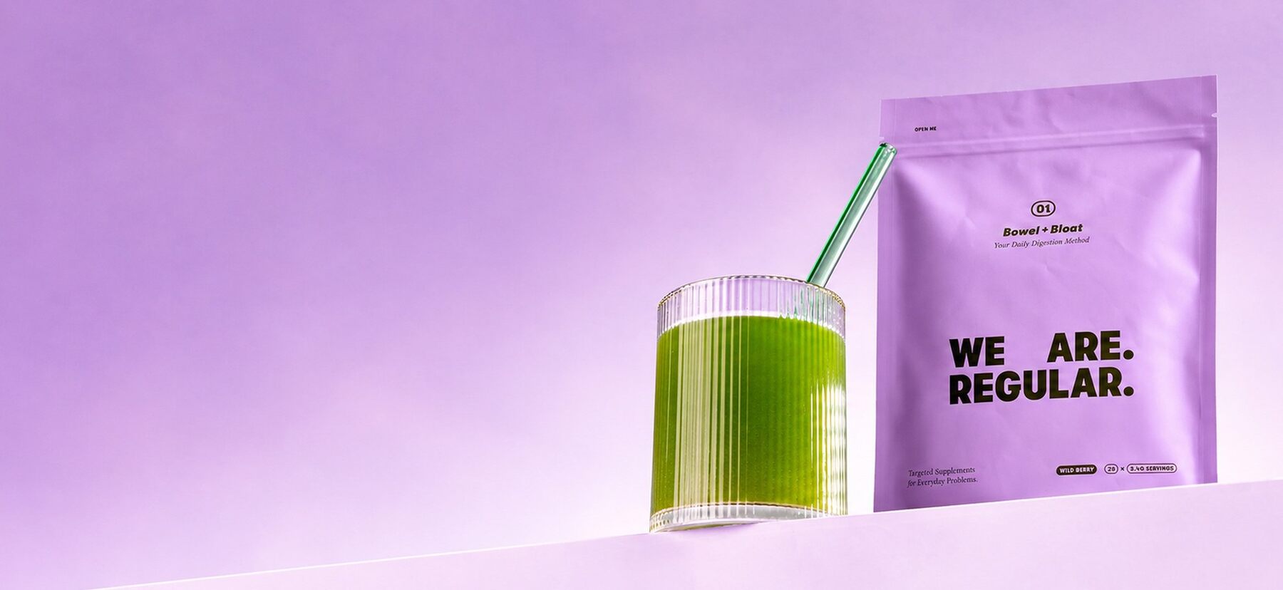 The We Are Regular pouch beside a glass of the mixed green drink with a straw